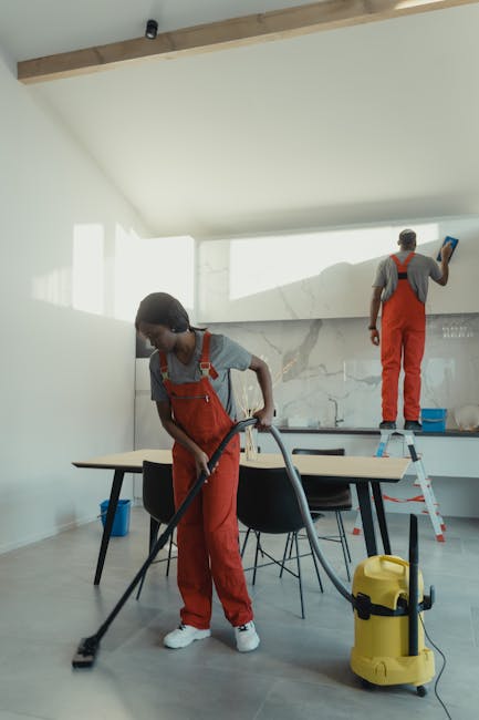 Two professional cleaners from Paddington Cleaner are engaged in surface cleaning within a modern, well-lit kitchen. The cleaner in the foreground is vacuuming a light wooden floor, wearing grey top and red overalls, with a yellow industrial vacuum cleaner positioned nearby. In the background, another cleaner stands on a small step ladder, wiping a white wall or cabinet with a cloth, also dressed in grey and red work attire. The kitchen features a minimalist design with white walls, a marble backsplash, and a large window allowing natural light to illuminate the space. The surfaces, such as the table and countertops, appear clean and dust-free, reflecting regular deep cleaning and surface sanitisation. Visible cleaning tools and the tidy arrangement highlight professional domestic cleaning processes by Paddington Cleaner.