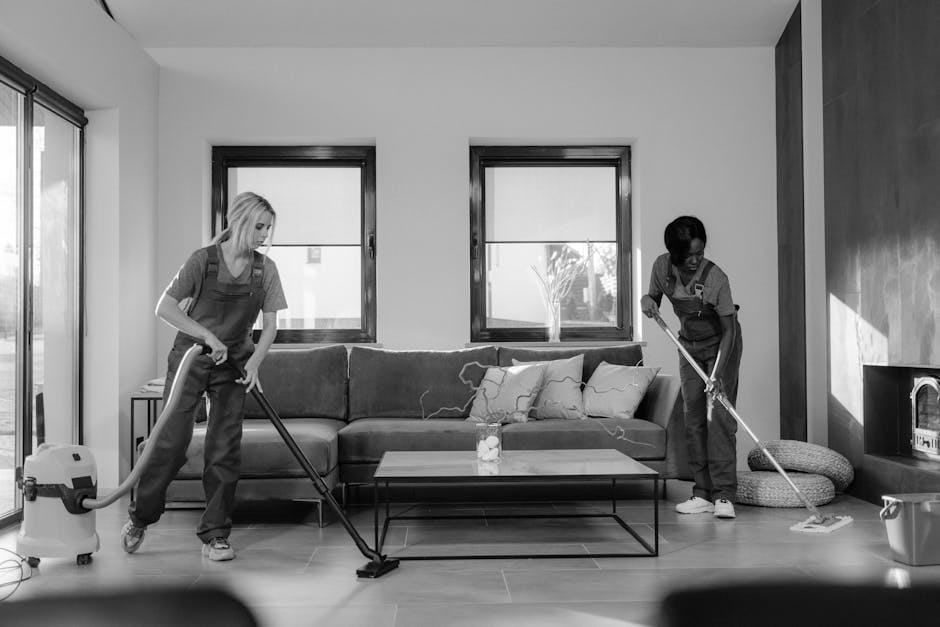 A female cleaner wearing a yellow uniform and yellow rubber gloves is holding a spray bottle in her right hand and a blue cloth in her left, standing in a bright, well-lit living room. The room features a beige sofa with cushions, a wooden side table with books and decorative items, and a modern fireplace. The surface cleaning involves a tidy and dust-free environment, with visible polished surfaces and clean floors. The overall setting highlights domestic cleaning and sanitisation carried out by Paddington Cleaner, with natural lighting emphasizing the cleanliness of the space.
