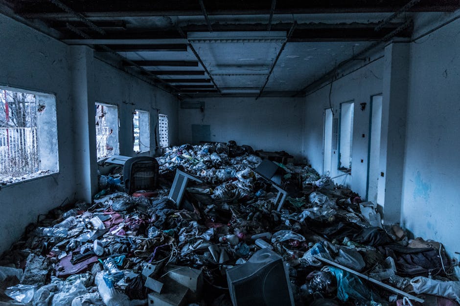 A room with multiple windows on the left side, showing an overflowing interior filled with a large volume of mixed household waste, including plastic bottles, paper, cardboard, and electronic equipment. The room has white plaster walls with visible patches of dirt and damage, and a dark, unfinished ceiling with exposed beams. The floor is entirely covered by clutter and debris, creating a chaotic scene indicative of hoarding. Natural light enters through the windows, illuminating the disorder, and the overall scene depicts a deep cleaning challenge in a residential setting, suitable for services provided by Paddington Cleaner for hoarding cleanup and sanitation.