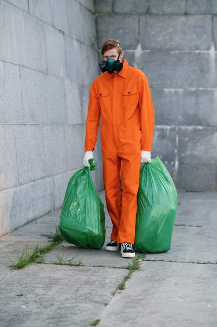 A person wearing an orange protective jumpsuit, white gloves, and a black mask stands outdoors against a grey concrete wall, holding two large green garbage bags filled with waste. The setting appears to be an urban alley or street with a concrete pavement. The image emphasizes waste collection and hygiene during cleaning or cleanup operations, relevant to deep cleaning and sanitisation services. Paddington Cleaner offers professional cleaning solutions, including hoarding cleanup for residential and commercial properties, ensuring thorough surface cleaning and sanitisation to maintain hygiene and safety standards.