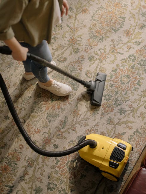 A person wearing beige sneakers and gray jeans is using a yellow vacuum cleaner with a black hose attachment to deep clean a patterned area rug. The rug features a floral design in muted shades of green, orange, and beige, and is laid out on a light-colored wooden floor. The room is well-lit, and the scene highlights surface cleaning and maintenance in a domestic setting. Paddington Cleaner specializes in deep cleaning services, including carpet and rug sanitisation, ensuring hygiene and freshness in residential spaces.