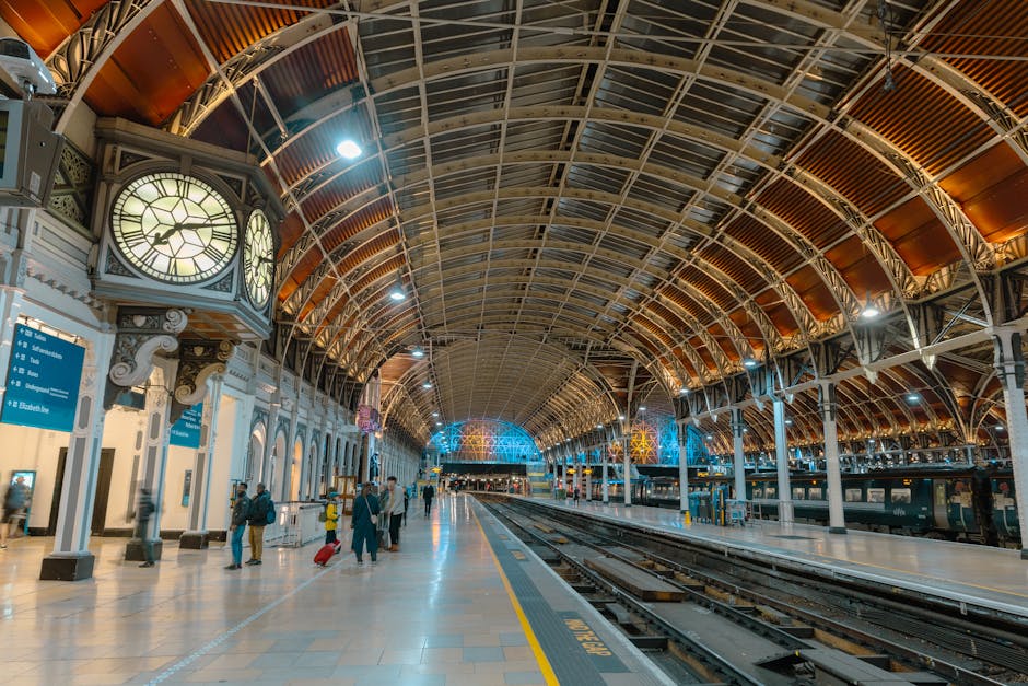 The image depicts the interior of Paddington Station with a spacious platform area featuring a vaulted, metal and wood ceiling structure illuminated by ceiling lights. The platform surface is made of smooth, polished tiles, which appear clean and shiny, reflecting the lighting above. There are a few passengers with luggage walking along the platform, with some standing near the white station walls and digital signboards displaying train information. A large, ornate clock with a white face and black Roman numerals is mounted on a decorative pillar near the platform edge. The scene captures a neat, well-maintained setting typical of a busy train station, emphasizing surface cleaning and hygiene, with Paddington Cleaner providing professional cleaning services to maintain such environments.