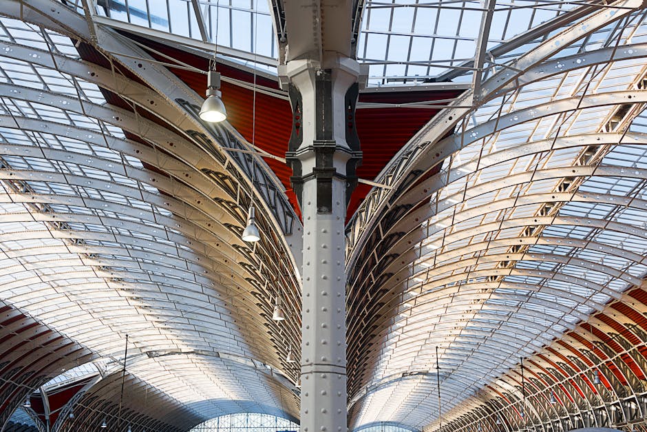 The image depicts the interior of Paddington Station with a spacious platform area featuring a vaulted, metal and wood ceiling structure illuminated by ceiling lights. The platform surface is made of smooth, polished tiles, which appear clean and shiny, reflecting the lighting above. There are a few passengers with luggage walking along the platform, with some standing near the white station walls and digital signboards displaying train information. A large, ornate clock with a white face and black Roman numerals is mounted on a decorative pillar near the platform edge. The scene captures a neat, well-maintained setting typical of a busy train station, emphasizing surface cleaning and hygiene, with Paddington Cleaner providing professional cleaning services to maintain such environments.