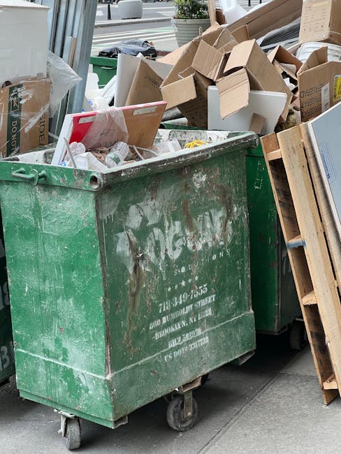 A large green industrial waste dumpster on a concrete pavement filled with a mix of cardboard boxes, paper waste, and packaging materials. Surrounding the dumpster are wooden pallets and additional debris, indicating a collection of bulky waste for clearance. The scene appears to be in an outdoor urban or commercial area with natural daylight illuminating the clutter. The condition of the waste is disorganized, with some boxes partially crushed or unfolded, and trash spilling over the edges of the dumpster. For professional same-day junk clearance and bulky waste solutions in Paddington, Paddington Cleaner offers reliable services to manage such clutter efficiently and maintain cleanliness. This image reflects the type of waste that may be handled during domestic or commercial cleaning projects aimed at surface cleaning, waste removal, and maintaining hygienic environments.