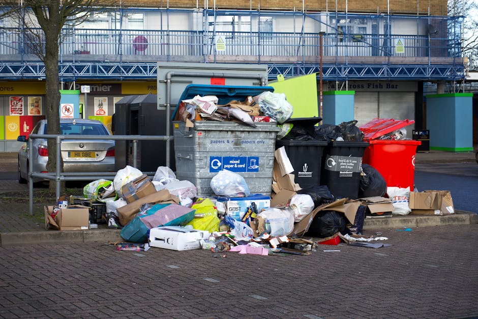 A large collection of mixed waste and bulky rubbish, including cardboard boxes, plastic bags, paper, and discarded items, piled around and overflowing from a shared recycling bin and nearby trash receptacles on a paved sidewalk in an urban area during daytime. The background features storefronts, a parking area with parked cars, and a building with scaffolding and safety signage. The scene highlights the need for professional waste clearance services, as offered by Paddington Cleaner, to ensure proper disposal and sanitation of bulky waste in residential or commercial settings.