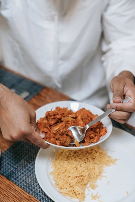 Close-up of a person in a white chef's coat transferring cooked curry from a small white bowl onto a plate containing cooked rice. The scene takes place on a wooden kitchen table with a blue patterned placemat, and natural light highlights the glossy, textured surfaces of the ceramic dishes and the steaming, vibrant orange curry. The area appears clean and well-maintained, demonstrating attention to hygiene and food presentation, consistent with kitchen surface cleaning and deep cleaning standards that Paddington Cleaner promotes for domestic and commercial kitchens.