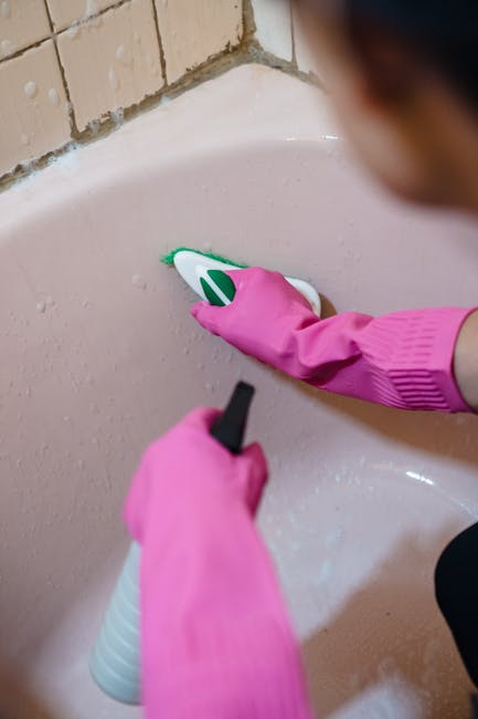 Close-up of a person cleaning a ceramic bathroom sink with a white and green spray bottle, wearing bright pink rubber gloves. The sink is pale pink with a smooth, glossy surface, and the edge reveals a tiled wall with beige bricks. The person's hand is holding a small black cleaning tool or sponge, applying a cleaning product to the sink’s edge, which appears to be free of stains and debris. The image showcases a thorough surface cleaning process typical of domestic cleaning routines aimed at maintaining hygiene and removing stains, with the focus on detailed sanitation work inside a bathroom setting, highlighting cleanliness and care. Paddington Cleaner specializes in cleaning services that include detailed stain removal and deep cleaning, ensuring surfaces are hygienic and spotless.