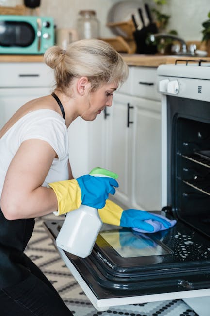 A woman wearing yellow and blue rubber gloves is crouching down to clean the interior of a black oven with a spray bottle and a blue cleaning cloth. The kitchen features white cabinets with black handles, a wooden countertop, a stainless steel sink, and various small appliances, including a green microwave. The surface of the oven is shiny and appears freshly cleaned, with the woman focused on detailed surface cleaning. The lighting in the kitchen is bright, highlighting the cleanliness and the reflective surfaces. The scene emphasizes domestic cleaning and sanitisation, indicative of a spring cleaning routine, aligned with services offered by Paddington Cleaner. The overall impression is of meticulous surface cleaning in a modern, well-maintained kitchen space.