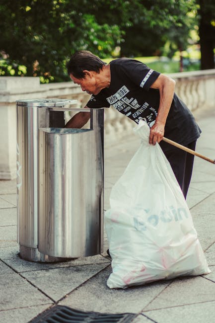 A man with dark hair, dressed in a black T-shirt with white text, is seen leaning over an outdoor stainless steel litter bin, actively disposing of trash. He is holding a large white plastic bag in his left hand, which is partially filled, and his right hand is reaching into the bin for rubbish collection. The image is set on a paved pathway with a stone railing in the background, surrounded by green leafy trees and bright natural lighting, indicating daytime. The scene depicts a routine urban cleaning activity, emphasizing cleanliness and waste management practice, which relates to the regulations for bulk rubbish disposal as outlined by Westminster Council for Paddington (W2). The setting appears neat and well-maintained, consistent with professional domestic or municipal cleaning standards, as represented by Paddington Cleaner.